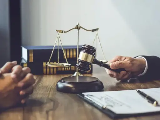 Dłoń trzymająca młotek sędziowski, waga sprawiedliwości i akta na drewnianym stole.