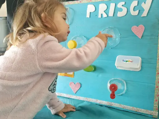 Bambina gioca con giochi motricità fine fai da te: un pannello azzurro con lettere glitterate, cuori rosa e oggetti da manipolare.