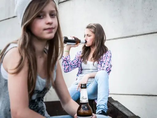Dwie nastolatki piją piwo, co może być początkiem alkoholizmu wśród młodzieży.