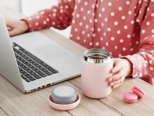 Kobieta przy laptopie trzyma kubek termiczny, obok leży nakrętka. Dowiedz się, jak wyczyścić nakrętkę od kubka termicznego, by napoje dłużej smakowały świeżo.
