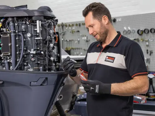 Un mécanicien effectue l'entretien d'un moteur hors-bord 4 temps Yamaha, tenant une bougie et un outil.