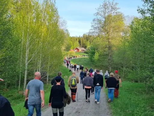 En grupp människor på en nattvandring Kinnekulle, på en grusväg genom en lummig skog. En röd stuga syns i bakgrunden.