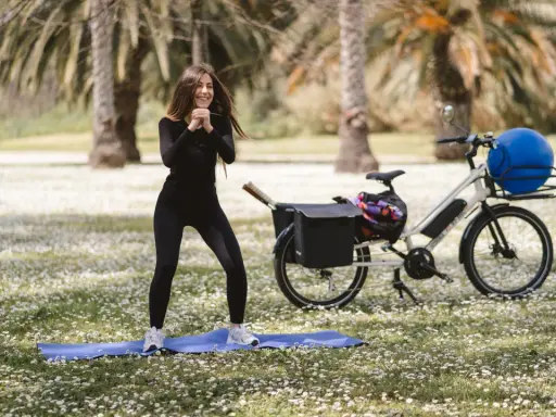 Donna sorridente fa esercizi all'aperto, accanto a una bicicletta per dimagrire con un grande pallone blu nel cestino.