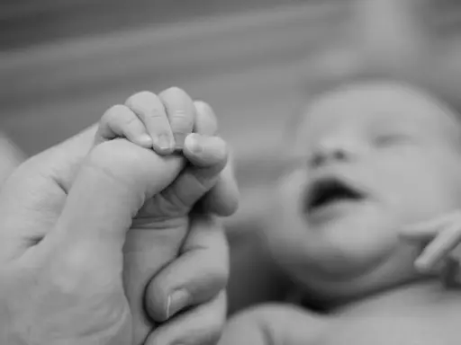 Dłoń dorosłego trzyma maleńką dłoń noworodka. Wiek postkoncepcyjny jest widoczny w delikatności paluszków.