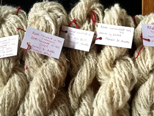 Buntar av ljusbeige garn, troligen mohair ull, med handskrivna etiketter som beskriver tvättmetoder och ursprung.