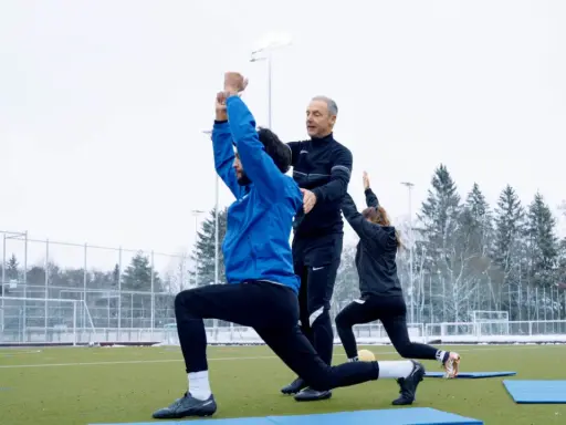 Athletik Training Fußball: Trainer korrigiert Ausfallschritt eines Spielers auf Kunstrasen. Zwei weitere Spieler machen Übungen im Hintergrund.