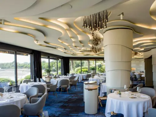 Salle élégante d'un restaurant chef de baie, tables dressées, vue sur la mer et jardin luxuriant. Plafond ondulé et luminaires modernes.