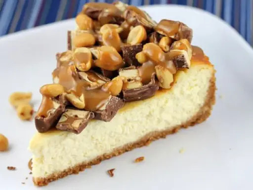 Kawałek sernika snickers z polewą karmelową i orzeszkami ziemnymi.