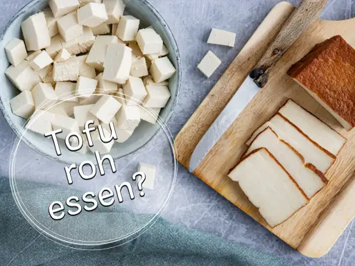 Würfel und Scheiben von Tofu liegen neben einem Messer auf einem Holzbrett. Die Frage "Kann man Tofu roh essen?" wird gestellt.