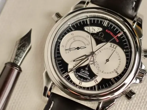 Elegante cronografo rattrapante Omega De Ville con quadrante nero e contatori argentati, accanto a una penna stilografica.