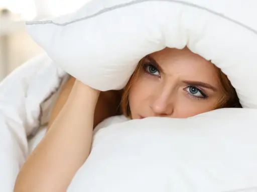Frau versteckt sich unter Kissen und Decke, weil sie nachts wach wird und Geräusche hört.