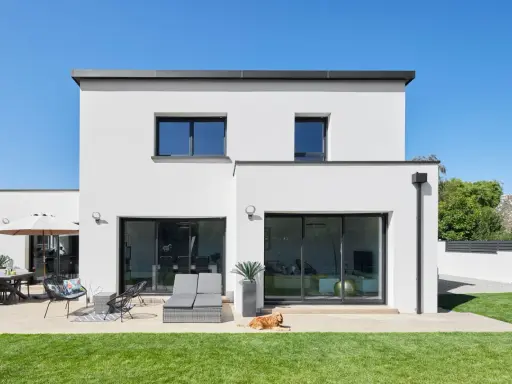 Maison moderne avec jardin, terrasse et chien. La ptz construction maison est visible sous un ciel bleu éclatant.