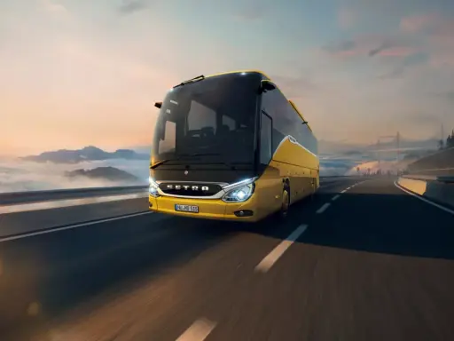 Autobús Setra amarillo brillante viaja por una carretera escénica al amanecer, con nubes y montañas de fondo.