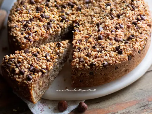 Una deliziosa torta di nocciole, ricoperta di granella di nocciole e gocce di cioccolato, con una fetta tagliata.