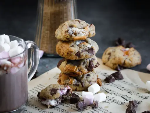 Un cookie chamallow fondant, empilé avec d'autres, à côté d'une tasse de chocolat chaud garnie de guimauves.