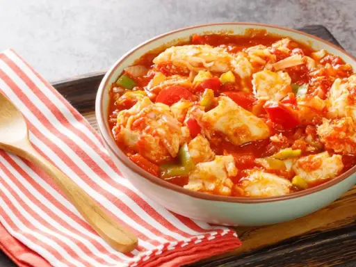 Un tazón de bacalao ajoarriero con pimientos y tomate, servido con una cuchara de madera sobre una servilleta a rayas.