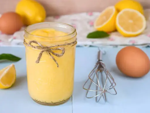 Un tarro de lemon curd casero, atado con cuerda, junto a limones, huevos y un batidor.