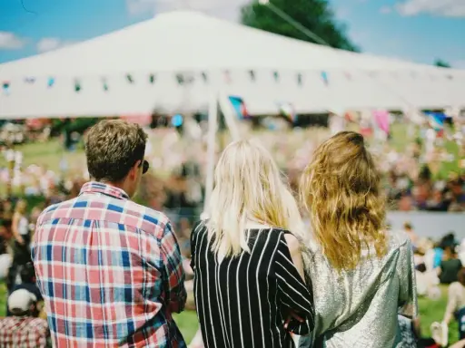 Tre vänner njuter av en solig dag på en musikfestival i Stockholm. En stor vit tältduk och flaggor syns i bakgrunden.