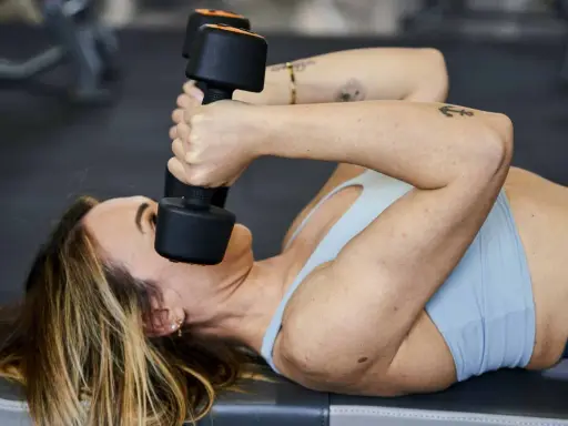 Donna sdraiata esegue skullcrushers con manubri, un esercizio efficace per i gruppi muscolari da allenare insieme, come tricipiti e spalle.