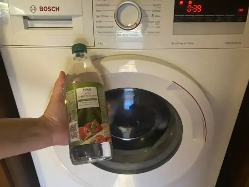 Ręka trzyma butelkę octu spirytusowego obok pralki Bosch. To początek domowego sposobu na mycie pralki octem.