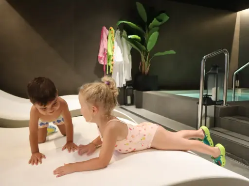 Niños jugando en un spa para niños, con una piscina al fondo y toallas colgadas.