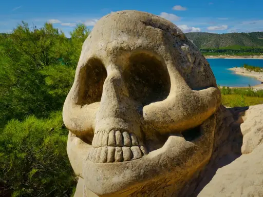 Escultura de calavera en la **ruta de las caras**, con un lago azul y colinas verdes al fondo.