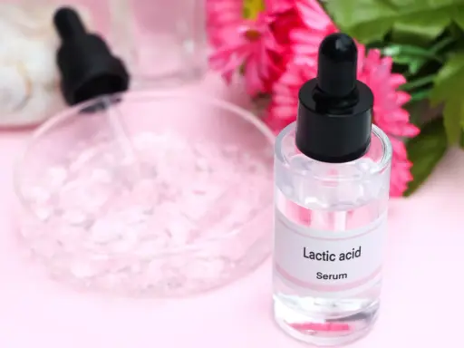 Serum z kwasem mlekowym w szklanej buteleczce z pipetą, obok różowych kwiatów i naczynia laboratoryjnego.