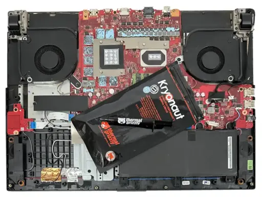 Wnętrze laptopa z wentylatorami i płytą główną, gotowe do wymiany pasty termoprzewodzącej. Opakowanie Kryonaut Thermal Grizzly leży na komponentach.