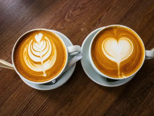 Zwei Cappuccinos mit Latte Art auf einem Holztisch. Sie bestehen aus Espresso, heißer Milch und Milchschaum.