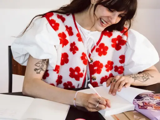 Une jeune femme souriante fait une dédicace livre, stylo à la main, devant un cahier ouvert et un paquet cadeau violet.