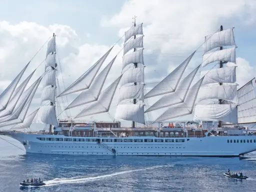 Ein majestätisches Segelschiff mit vollen weißen Segeln, das wie ein **sea cloud spirit** über das blaue Meer gleitet. Kleine Beiboote sind in der Nähe.