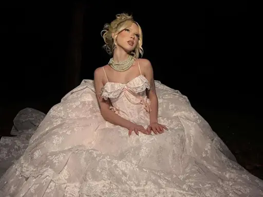 A woman in a voluminous, lace-trimmed gown and pearl necklace embodies modern romantic style, posing in a dark, outdoor setting.