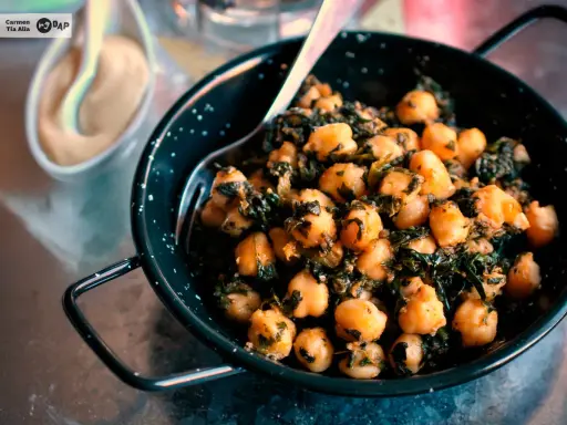 Un plato de garbanzos con espinacas, servido en una sartén negra con un tenedor. Al fondo, una pequeña taza con una salsa cremosa.