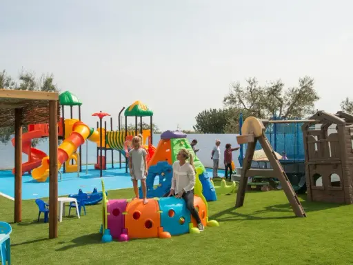 Un parco giochi colorato in una masseria per bambini in Puglia, con scivoli, tunnel e altalene. Famiglie si divertono sotto il sole.