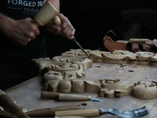Händer formar träsnideri med mejsel och klubba. Det ser ut som att träet är mjukt som lera, perfekt för detaljerat arbete.