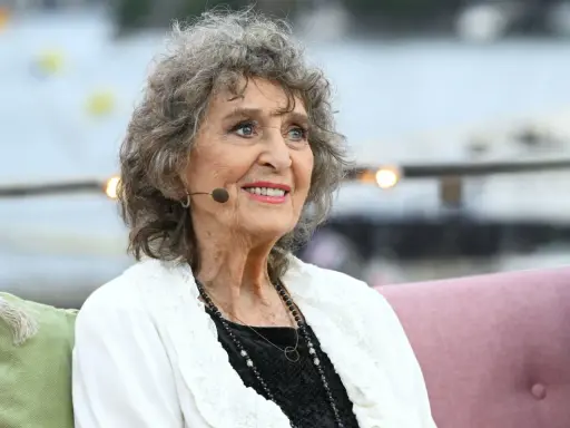 Siv Malmkvist, med sitt karaktäristiska lockiga gråa hår och ett varmt leende, sitter och tittar åt sidan. Hon bär en vit kofta över en svart klänning och smycken.