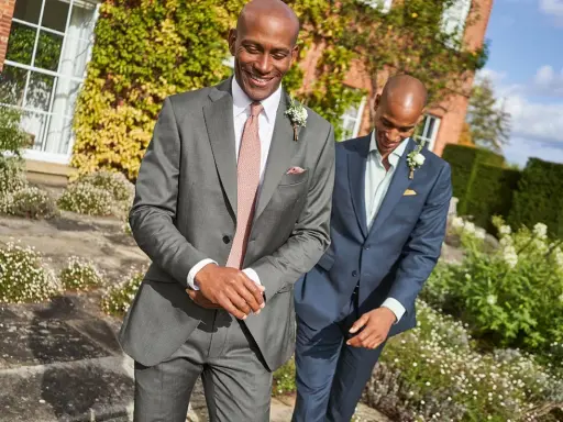 Two men in suits walk outdoors. The question "do you have to wear a suit to a wedding?" is answered by their stylish attire.