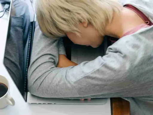 Zmęczona osoba śpi na laptopie. Czy leczenie odwykowe a zwolnienie z pracy to trudny wybór? Kawa obok.