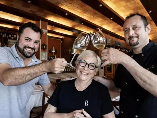 Dos hombres y una mujer brindan con copas de vino en Ca la Pepa, un restaurante acogedor con estanterías de vino.