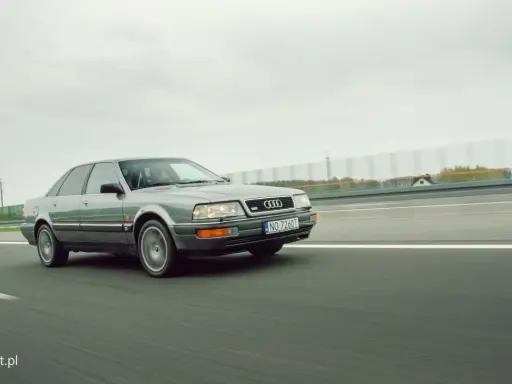 Szary Audi V8 pędzi po autostradzie, chmury na niebie i zielone drzewa w tle.