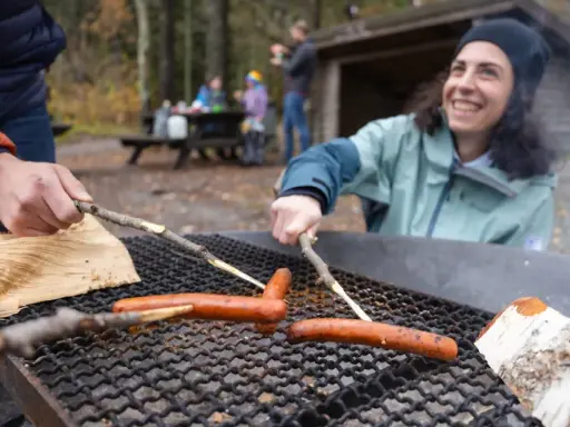 Vänner grillar korv vid en eldstad på Otto Pers gård. En kvinna ler glatt medan hon grillar.