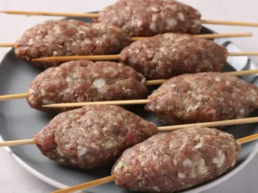 Siedem surowych kofta kebab na patyczkach, ułożonych na szarym talerzu, gotowych do grillowania.
