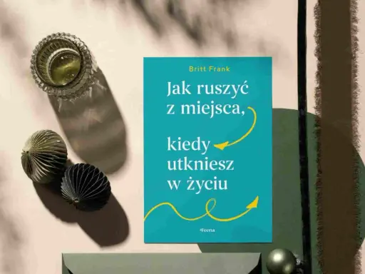 Książka "Jak ruszyć z miejsca, kiedy utkniesz w życiu" Britt Frank. Obok szklanka z napojem i ozdobne kule. Idealna lektura, gdy dopada lenistwo.