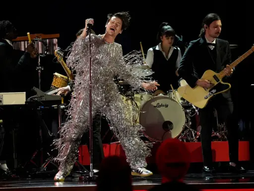 Harry Styles i glittrig outfit på en magisk harry styles konsert. Bandet spelar bakom honom, en gitarrist i kostym och en trummis.