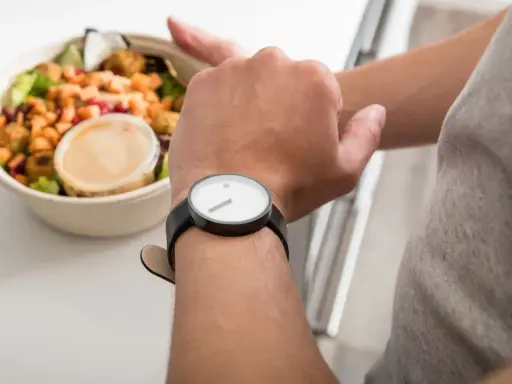 Mann schaut auf seine Uhr, neben einer Schüssel mit Salat. Die Frage: darf man fasten wenn man krank ist?