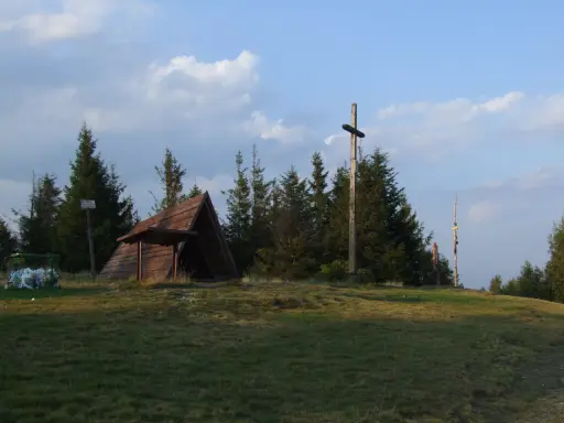 Drewniana wiata, krzyż i kosz na śmieci na tle drzew i nieba.