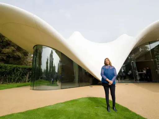 Zaha Hadid steht vor einem ihrer ikonischen zaha hadid bauwerke, einem geschwungenen, weißen Dach mit Glasfront.