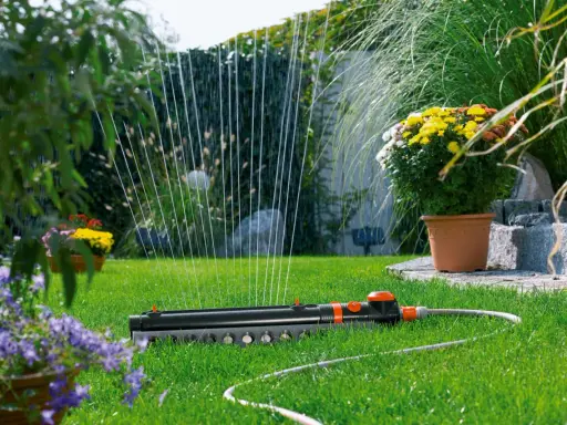 Sistema de riego automático Gardena en césped verde, con flores amarillas y moradas al fondo.