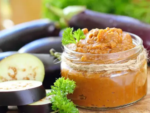 Un bocal de le vrai caviar d'aubergine, garni de persil frais, avec des aubergines entières et tranchées en arrière-plan.