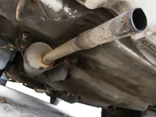 Vista inferior de un coche, mostrando el sistema de escape con un silenciador y tuberías oxidadas. El colector de escape es una parte crucial del sistema.